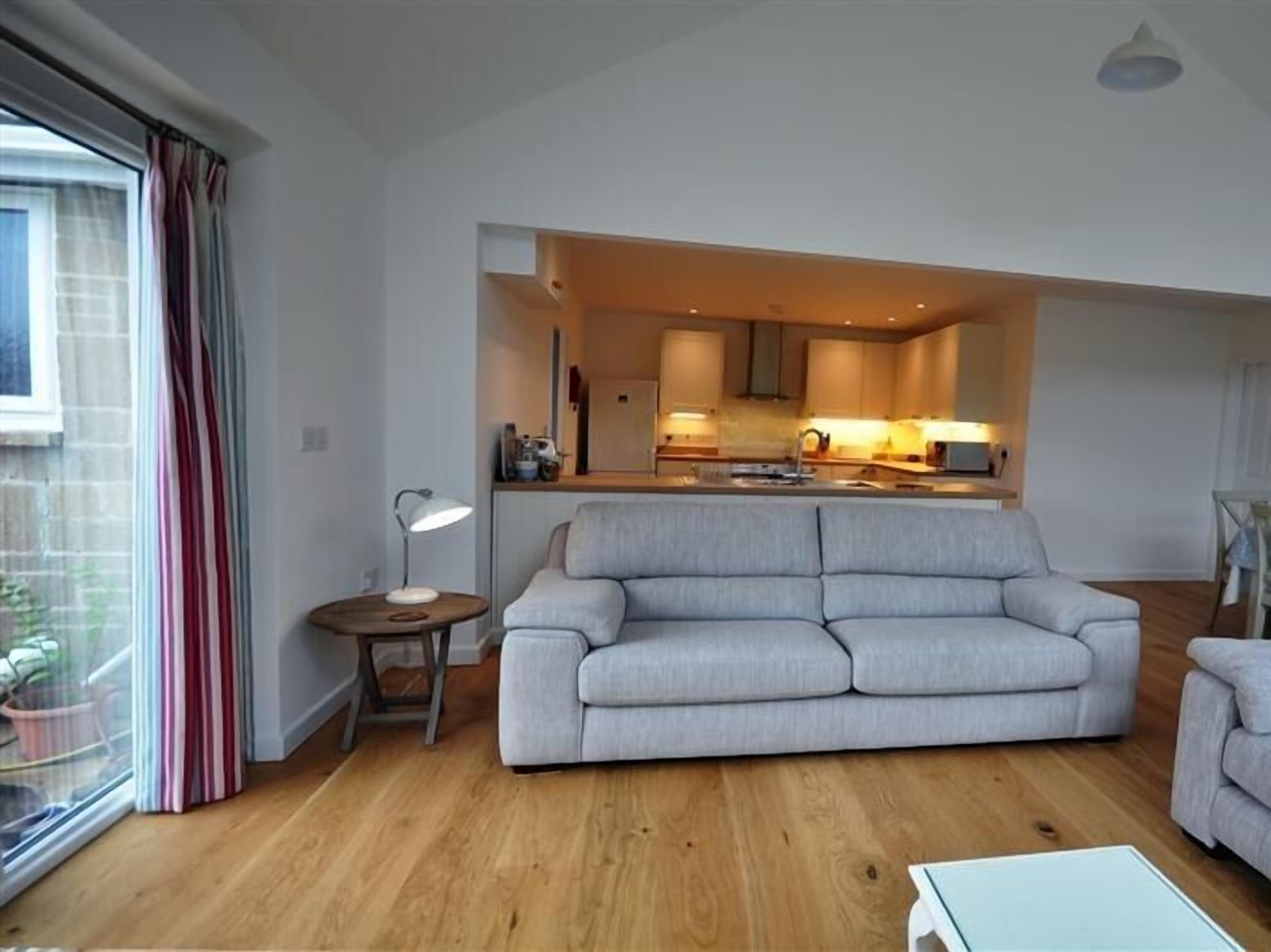 A modern living room with a grey sofa, a small round table with a lamp, and a kitchen in the background.