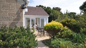 Una casa in pietra con tetto in tegole, un portico coperto con tavolo e sedie, e un giardino curato con arbusti e un grande cespuglio.