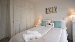 Una camera da letto con un letto grande, tende bianche, un quadro con case sulla spiaggia e un comodino con una lampada.