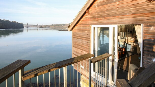 Une cabane en bois avec une porte en verre donnant sur un intérieur aménagé, et offrant une vue sur une rivière paisible, avec un pont visible au loin.