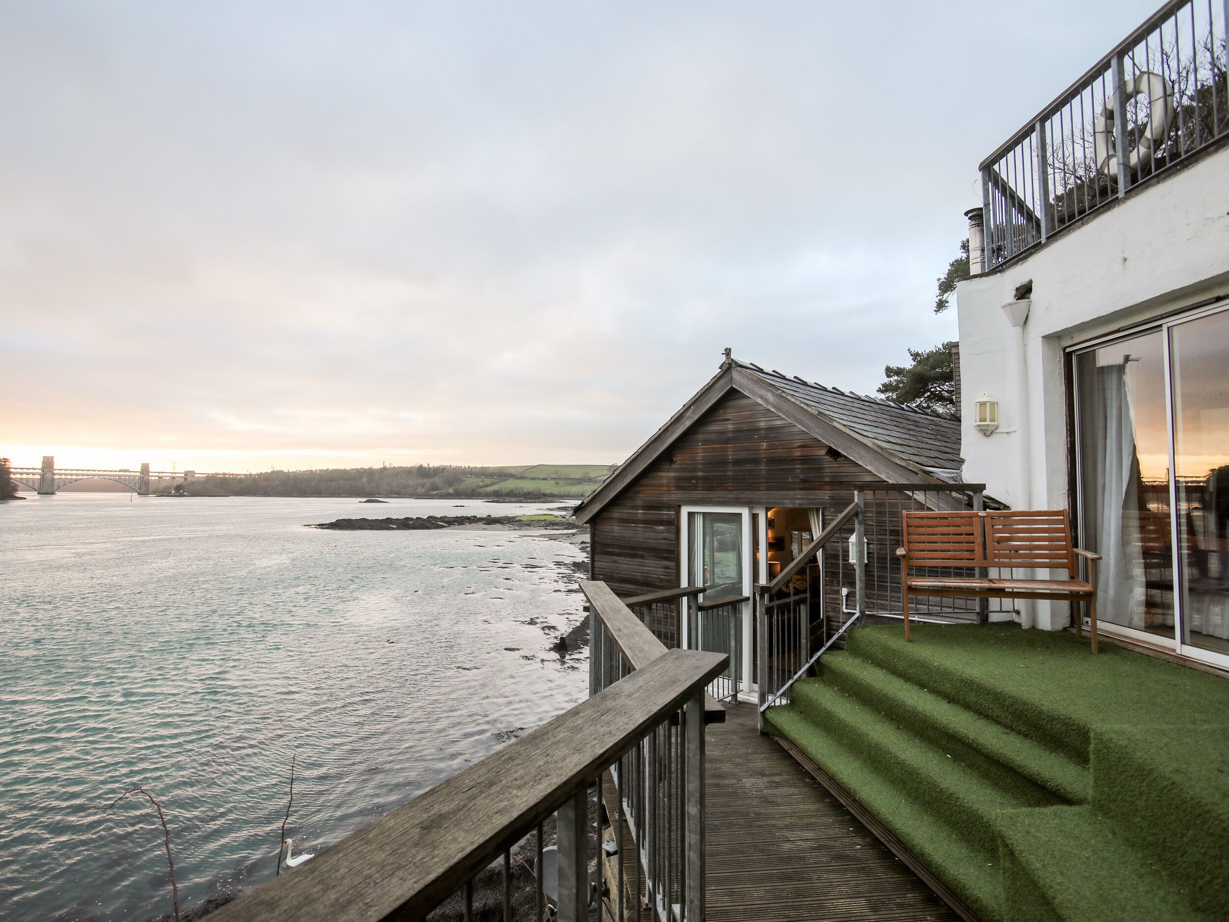 A wooden cabin with a glass door, a wooden deck, and a view of a river and bridge.