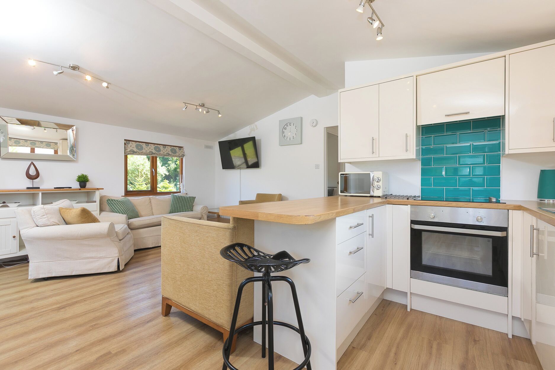 A modern kitchen with white cabinets, a wooden countertop, and a built-in oven. There is a small island with a bar stool, and a TV mounted on the wall.