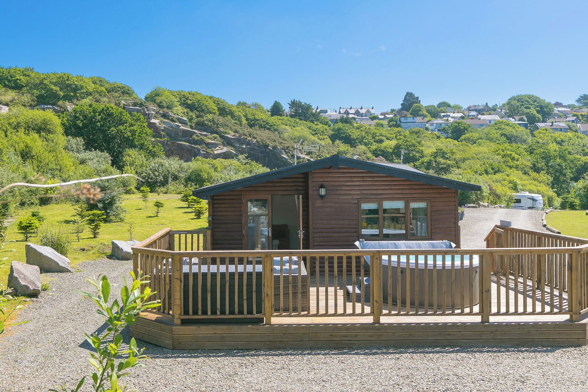 A wooden cabin with a deck, surrounded by greenery and hills.