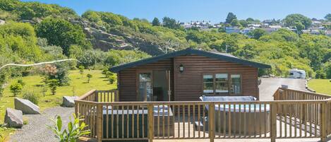 A wooden cabin with a deck, surrounded by greenery and hills.