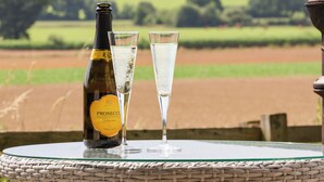 A bottle of Prosecco and two glasses on a round table with a view of a green field and trees in the background. - Bellwether (York)