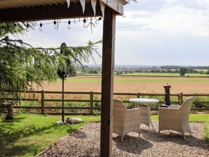 A covered outdoor seating area with wicker chairs and a small table, overlooking a rural landscape with fields and a wooden fence.