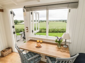 A kitchen with a wooden table, two chairs, a window with a view of a green field, and a lamp on the table.