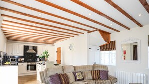 Un vaste espace de vie avec un grand canapé en forme de L, des poutres en bois au plafond, une cuisine à l’arrière-plan et une vue sur l’extérieur par une fenêtre.