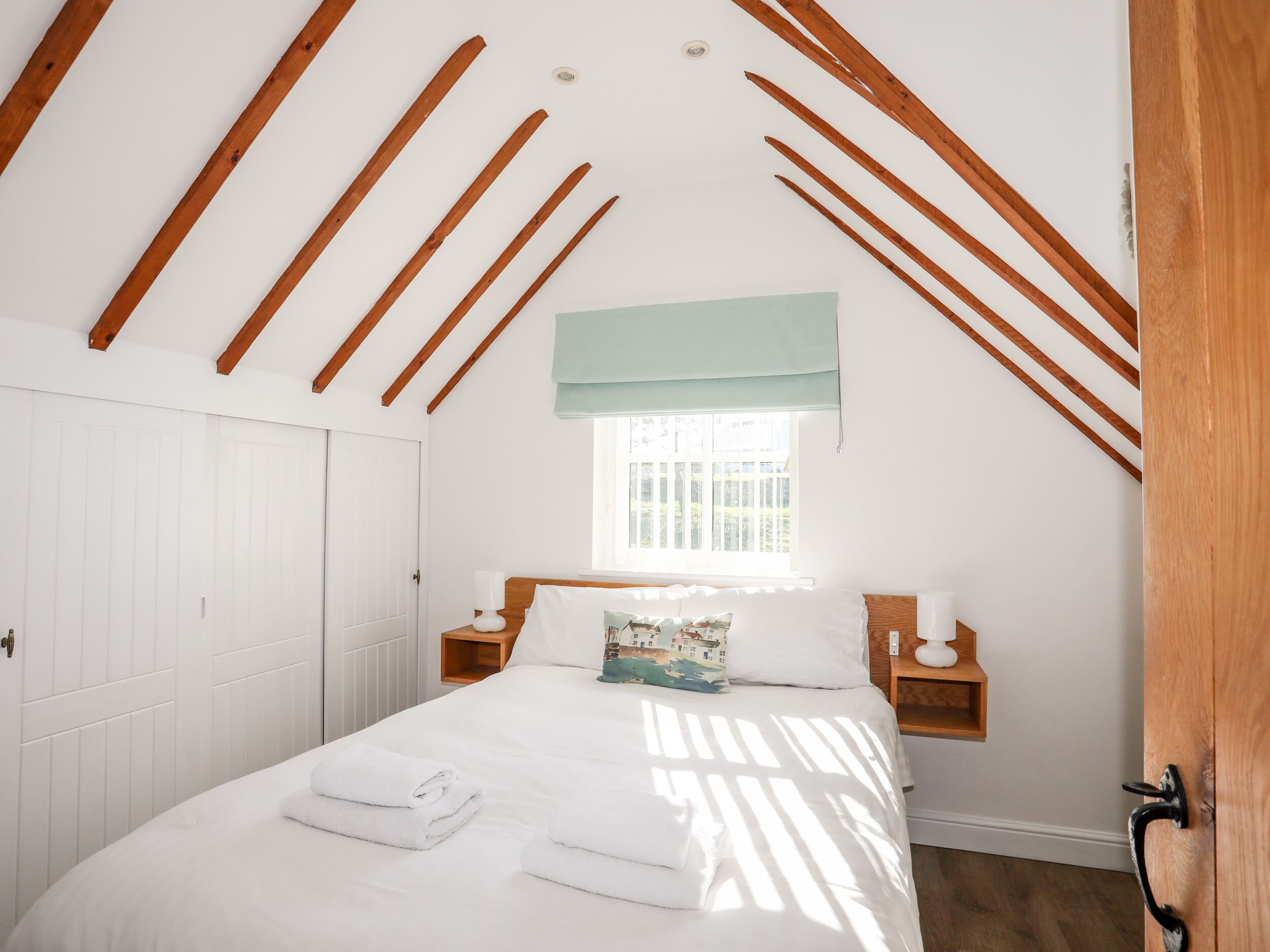 A bedroom with a sloped ceiling, a window with a light blue valance, a bed with white linens and a decorative pillow, and a bedside table with a lamp.
