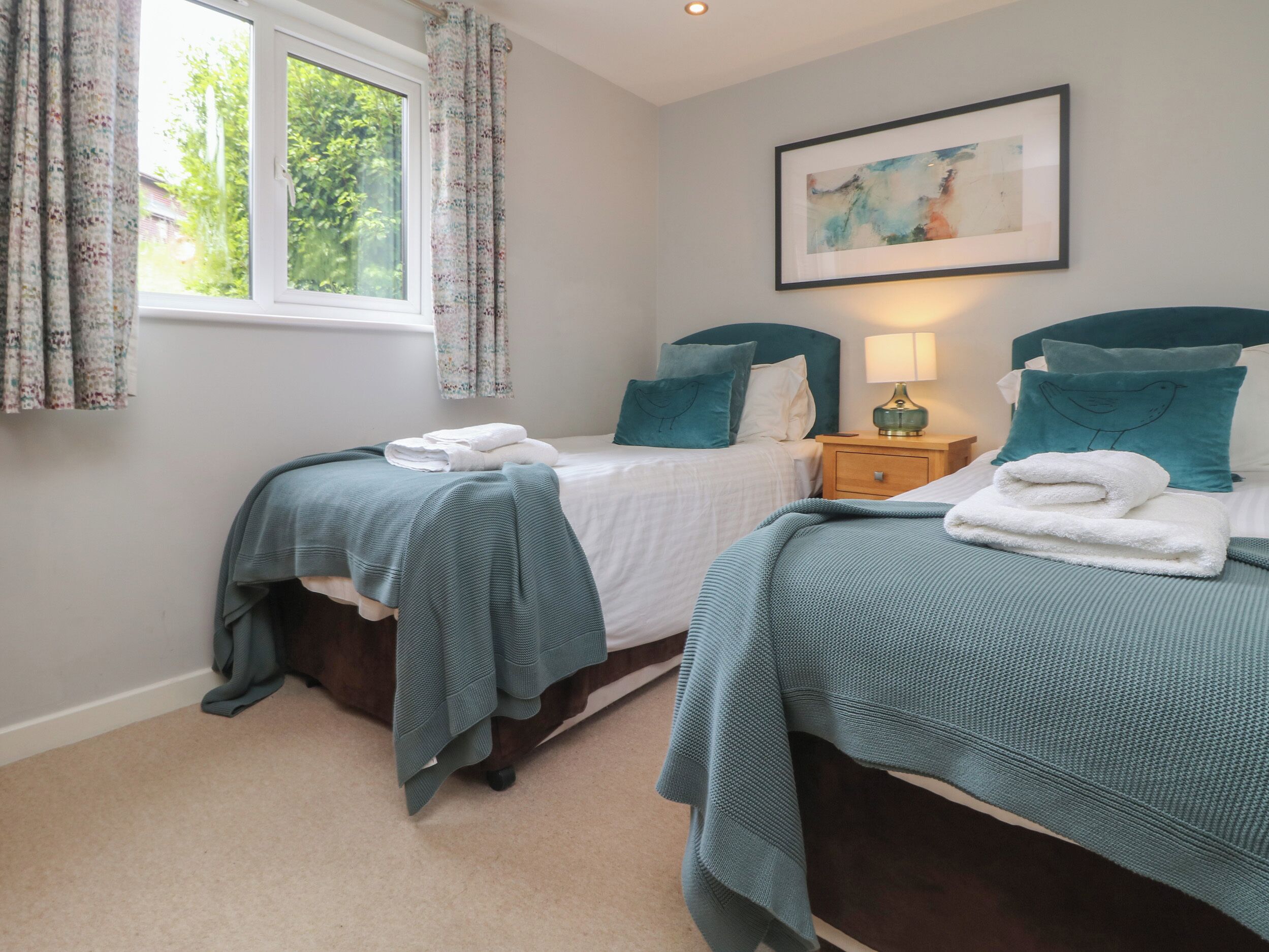 A bedroom with two beds, each with teal bedding and pillows, a nightstand with a lamp, and a framed picture on the wall.