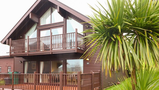 Une cabane en bois à deux étages, dotée d’un balcon, d’une allée en gravier et d’un palmier au premier plan.