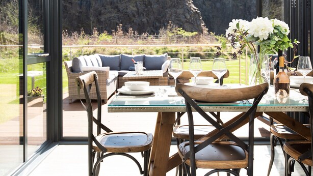 A dining area with a glass table, wooden chairs, and a sofa with cushions. There is a vase with flowers on the table. - Lambs Lodge (Bamburgh)