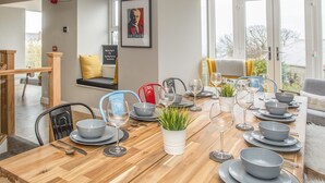 A dining area with a wooden table set for six, surrounded by chairs, and a view of the outdoors through large windows. - Cambrian House (Criccieth)