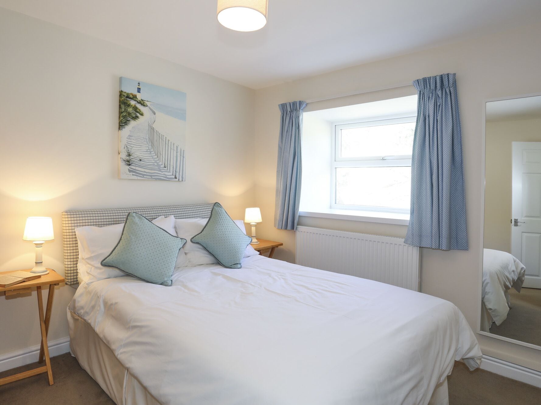 A neatly arranged bedroom with a bed, two bedside tables, a window with curtains, a wall-mounted artwork, and a mirror.