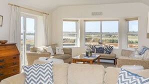 Ein geräumiges Wohnzimmer mit einer großen Couch, zwei Sesseln und einem Couchtisch. Mehrere Fenster bieten Ausblick ins Freie.