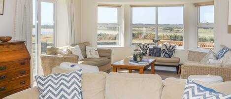 A spacious living room with a large sofa, two armchairs, and a coffee table. There are several windows providing views of the outdoors.