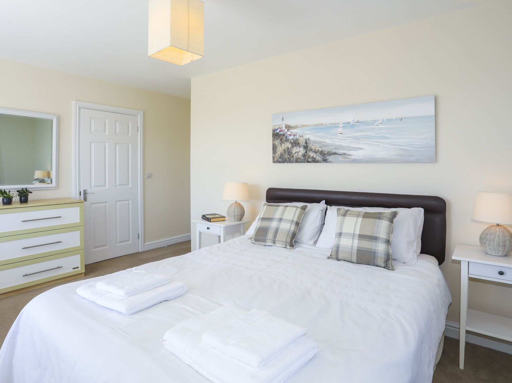 A neatly made bed with white linens and pillows, a bedside table with a lamp, a painting on the wall, and a dresser with a mirror.