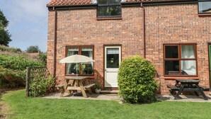 A brick house with a tiled roof, two windows with curtains, a door, and a small garden with a wooden table and chairs. - Fauna's Corner (Whitby)