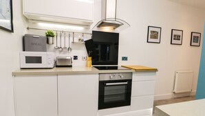 Une cuisine moderne avec des meubles blancs, un four encastré, un four à micro-ondes et un grille-pain. On y trouve des ustensiles suspendus et une petite plante posée sur le plan de travail.