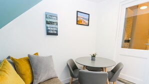 A cozy room with a grey sofa, yellow and grey cushions, a small round table with two chairs, and a potted plant. There are two wall decorations, one with text and another with a landscape painting.