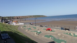 A coastal area with a sandy beach, a golf course, and a grassy slope.