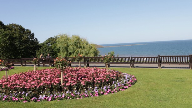 Un jardin aménagé avec une grande variété de fleurs et un espace agrémenté de bancs, offrant une vue sur un plan d’eau.