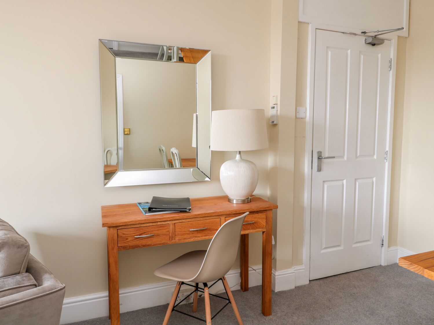 A wooden desk with a chair, a white lamp, and a large mirror on the wall.
