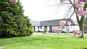 A white building with a grey roof, surrounded by a well-maintained lawn and flowering trees. - Orchard Cottage (Shaftesbury)