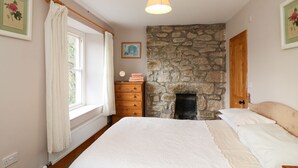 A bedroom with a stone fireplace, a wooden dresser, a bed with white bedding, and a window with curtains. - Brimpts Cottage (Yelverton)