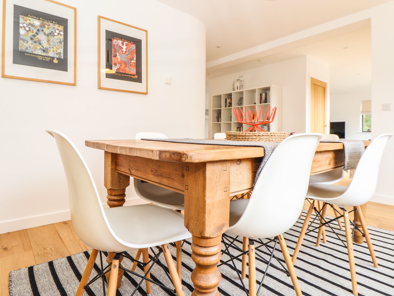 A dining area with a wooden table, white chairs, and a striped rug. There are framed artworks on the wall and a bookshelf in the background.