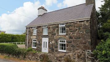 A stone house with a white door and multiple windows, surrounded by a fence and greenery.