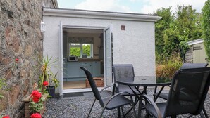 A small outdoor kitchen with a dining table and chairs, a stone wall, and a garden with red flowers. - Tan Y Goeden (Pwllheli)