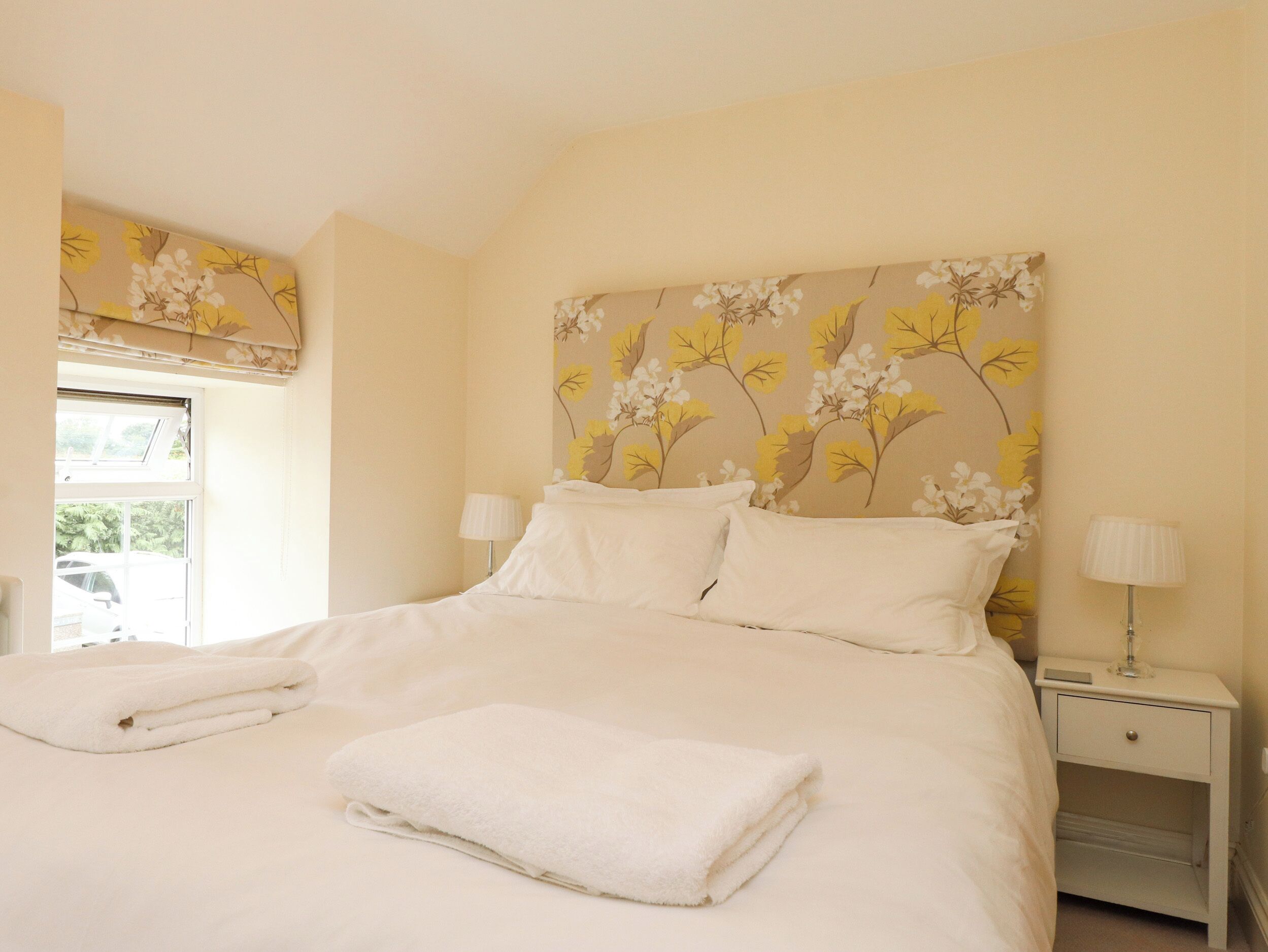 A neatly made bed with white linens and a folded white towel, a bedside lamp, and a window with a view of greenery.