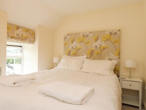 Un lit bien fait, avec du linge de lit blanc et une serviette blanche pliée, une lampe de chevet et une fenêtre donnant sur un paysage verdoyant.
