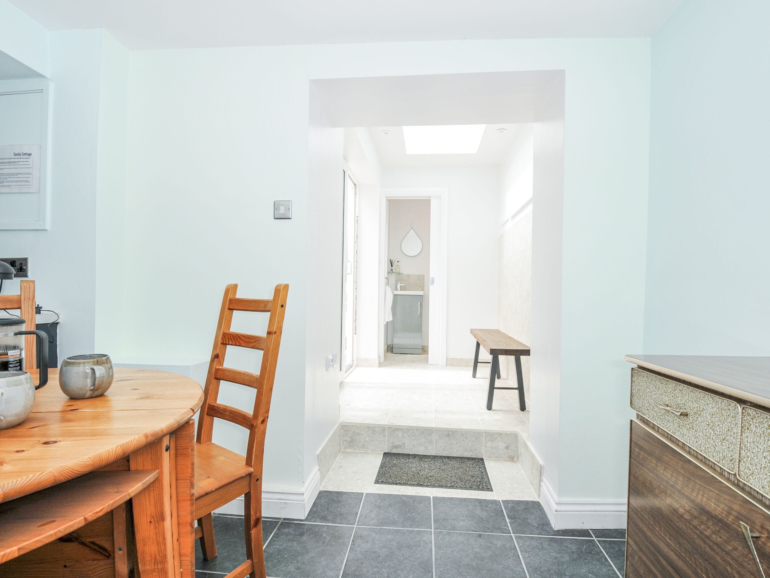 A modern kitchen with a wooden dining table and chairs, a built-in oven, and a view into the adjacent bathroom.