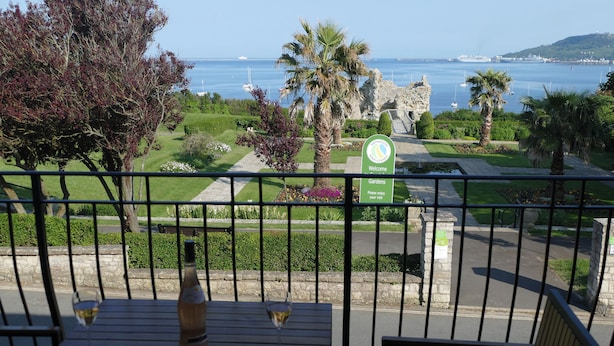 Ein Balkon mit Tisch, zwei Weingläsern und einer Flasche. Im Hintergrund ein Schild für die Gärten und der Blick auf das Meer mit Booten.