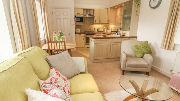 A living room with a green sofa, a glass coffee table, and a kitchen in the background. - Apple Tree Cottage (Windermere)