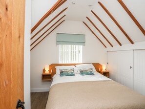 A bedroom with a sloped ceiling, a bed with a beige bedspread and decorative pillows, two bedside lamps, a window with blinds, and a door with a handle.