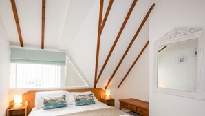 A bedroom with a sloped ceiling, wooden beams, a bed with a beige quilt, two pillows with nautical prints, a bedside lamp, a nightstand, a wooden dresser, and a mirror with a floral frame. - Tyn Towyn - Ty Glas Golau (Holyhead)