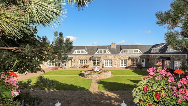 A stone building with a well-maintained lawn, a fountain, and a variety of flowers. - Tyn Towyn - Ty Glas Golau (Holyhead)