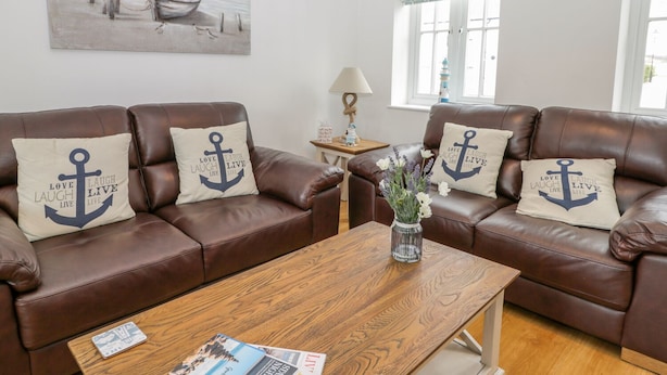 A living room with two brown leather sofas, each with a pillow featuring an anchor and the phrase "Love Laugh Live." A wooden coffee table is in the center, and there's a painting of a boat on the wall. - Bayside Cottage (Filey)