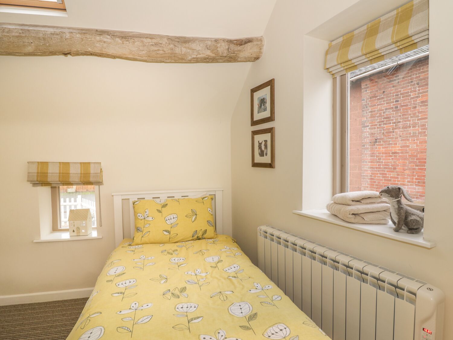 A bedroom with a yellow floral bedspread, a window with a view of a brick building, a radiator, and a small shelf with towels.