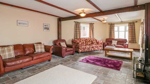 A spacious living room with a brown leather sofa, a patterned sofa, and a red leather armchair. There is a wooden coffee table and a television on a stand. The room has a tiled floor and wooden beams on the ceiling. - 5 Cliff Lane (Bridlington)