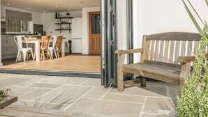 An open patio with a wooden bench, a dining table with chairs, and a kitchen visible through the open sliding doors. - The Beach Halt (Perranporth)