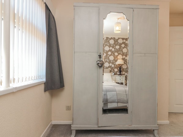 A room with a grey wardrobe, a window with blinds, and a mirror reflecting a bed with a floral wallpaper.