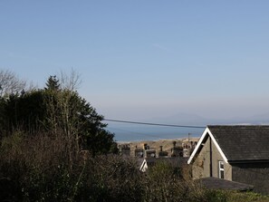 A coastal house with a stone chimney, surrounded by dense vegetation and a clear view of the sea and distant hills.