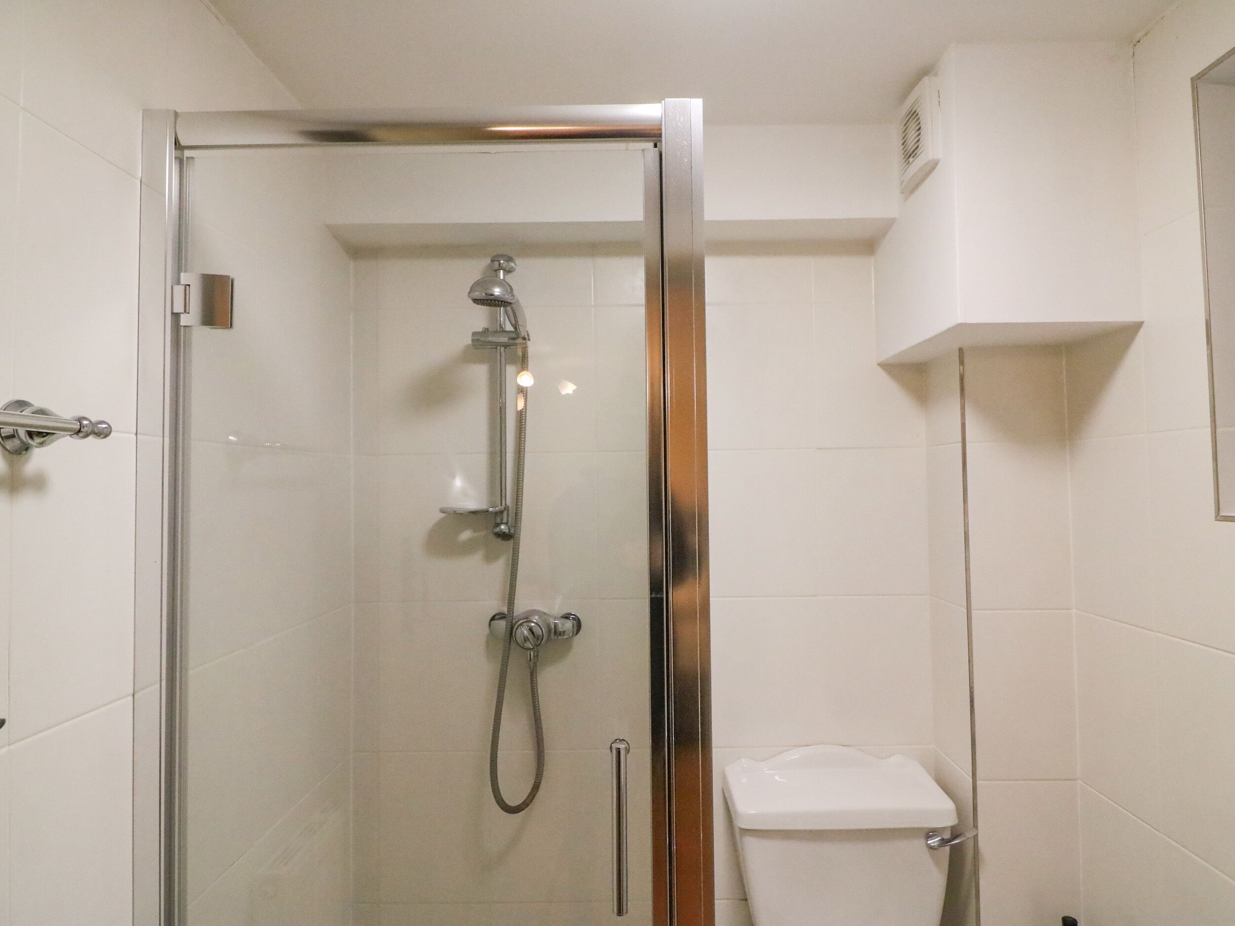 Cabina doccia con pareti in vetro, dotata di soffione manuale e doccetta, e water con pulsante di scarico.
