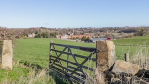 A rural landscape with a wooden gate, stone pillars, and a distant view of a village with buildings and a church. - Raygill Cottage (Whitby)