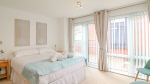 Quarto com cama, mesa de cabeceira, cadeira e uma janela grande com cortinas.