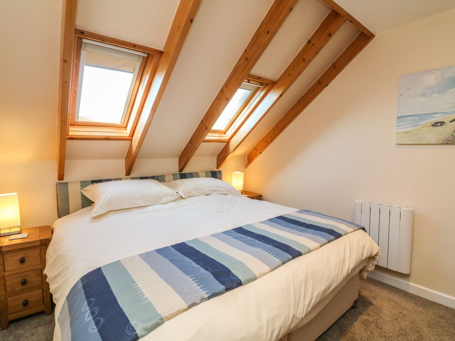 Ein Schlafzimmer mit schrägen Wänden, einem Bett mit gestreifter Bettdecke, einem Nachttisch mit Lampe und einem Bild an der Wand.